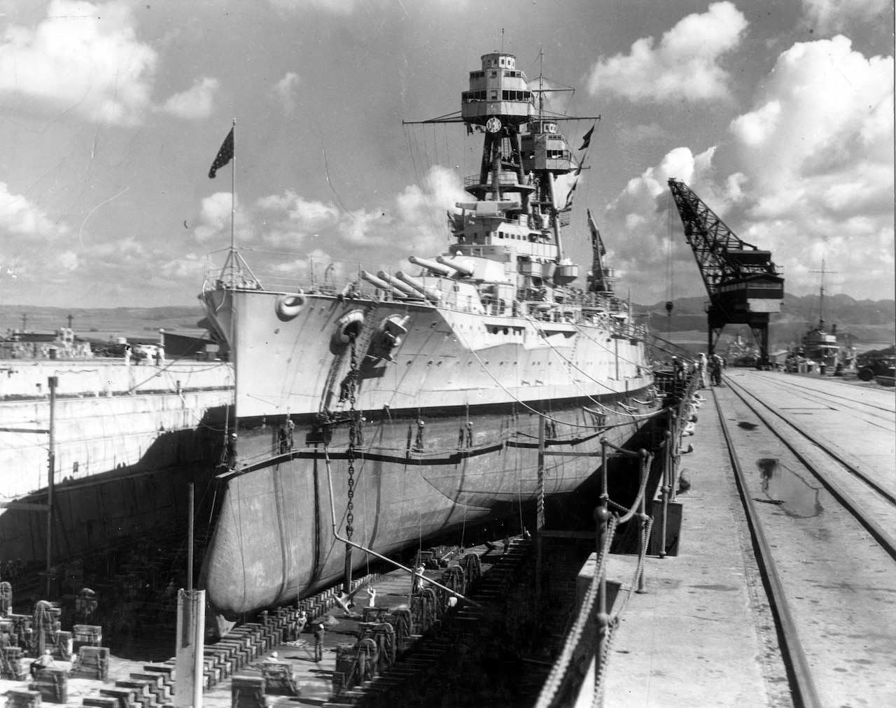 In drydock a battleship undergoes repairs during a partly cloudy day.