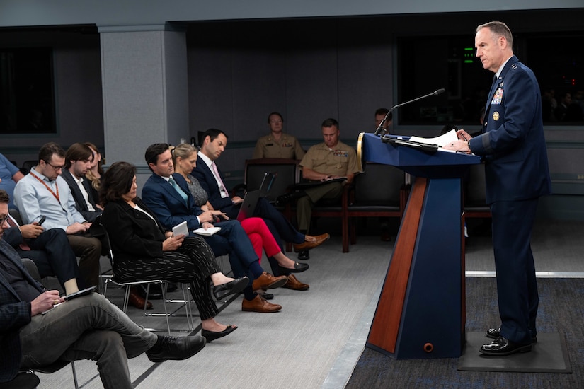 A military officer speaks from a lectern with the reporters seated in a room.