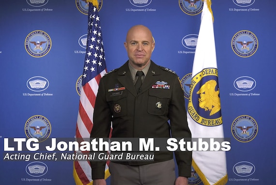 A man in a dress military uniform stands looking into the camera. Behind him is the United States flag to the left and the flag of the National Guard Bureau to the right.