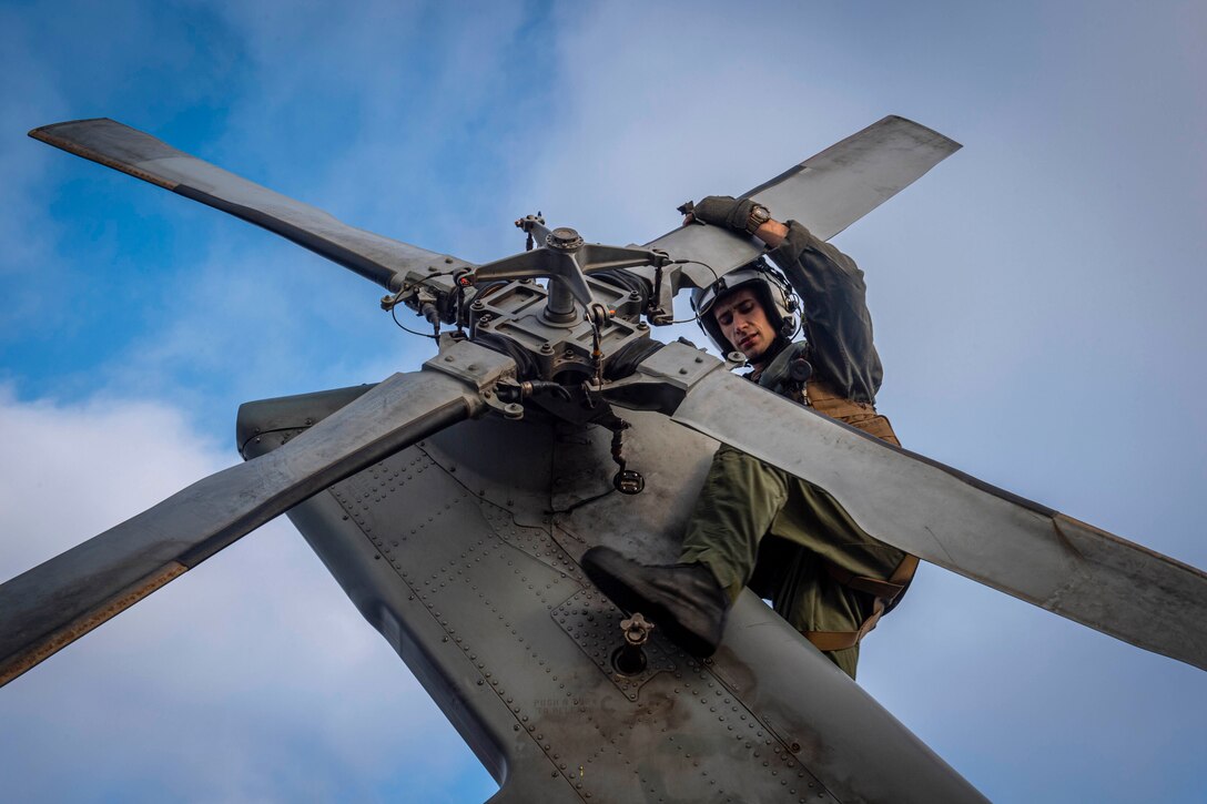 A sailor holds on to a propeller blade while perched on the frame of a helicopter at daylight.