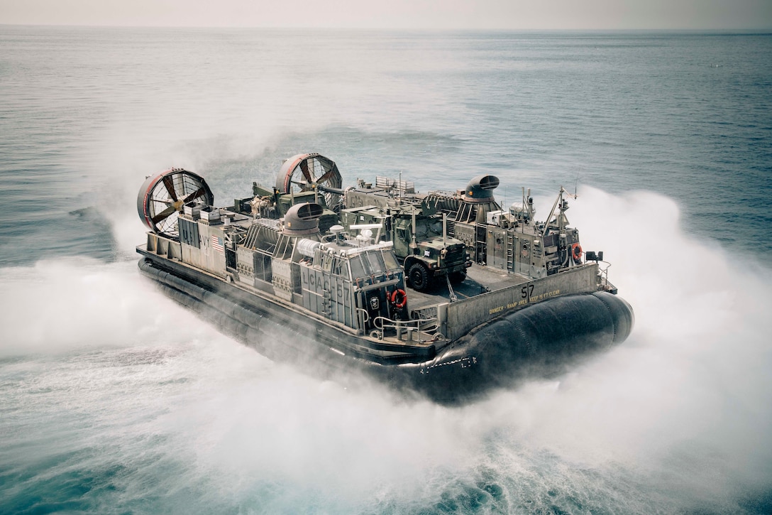 A military air-cushioned landing craft operates in open water during daylight creating a wake.