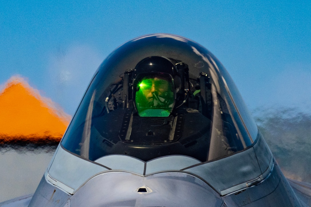 A close-up of an airman wearing a helmet illuminated by a small green light sitting in a cockpit.