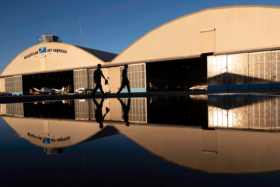 The silhouette of two airmen are seen walking in front of an airport with open hangars as their reflection is shown on ground.