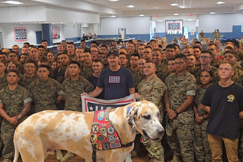 A large group of people poses for a photo while one holds a USO flag. A large dog stands in front of them.