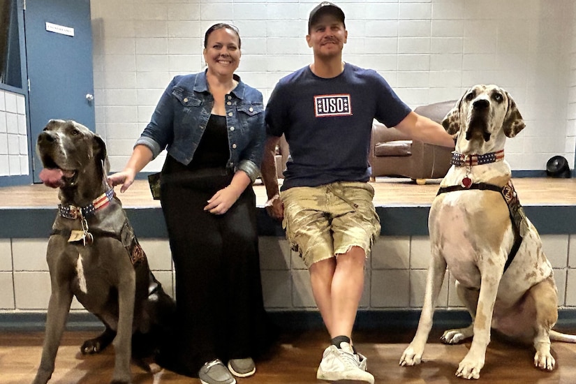 Two people lean on a stage while petting two large dogs who are sitting on the floor.