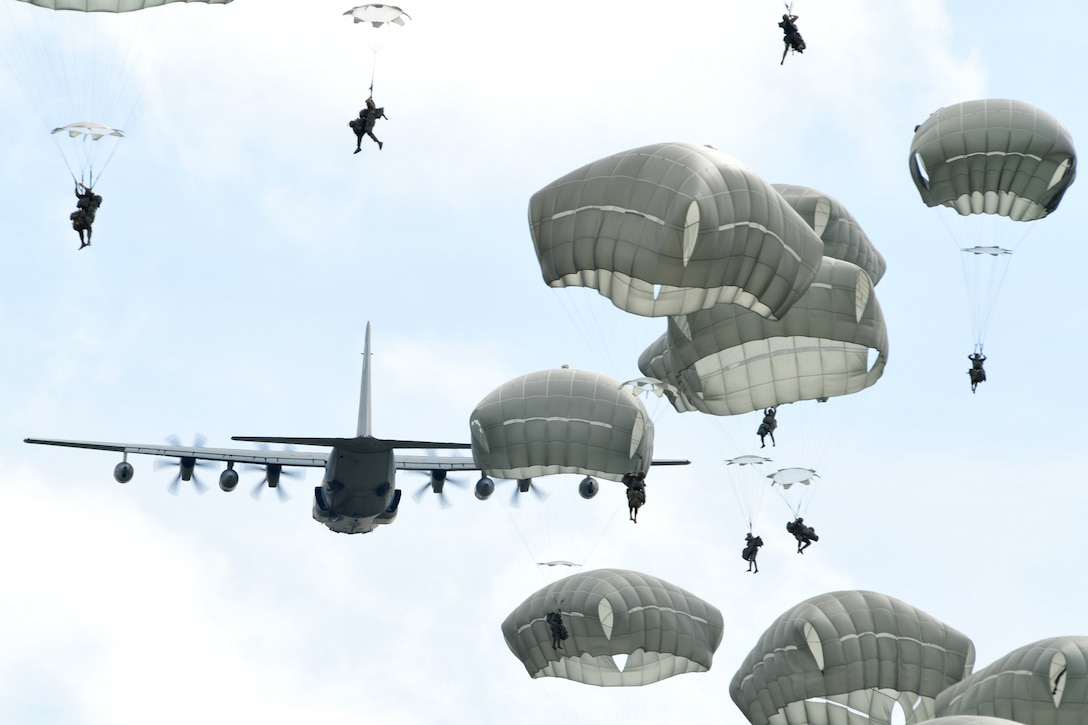 Parachutes and paratroopers fill the sky while a large aircraft flies away from the camera in the background.