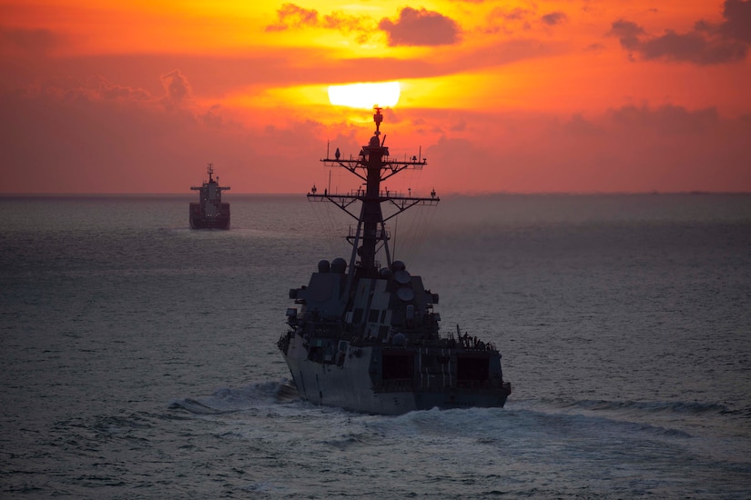 Two military ships steam through the water as the sun sits above the horizon creating a yellow and orange sky.