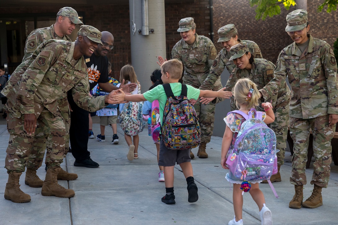 Smiling airmen stand in two lines while giving high-fives and fist bumps to children walking between them into a school.