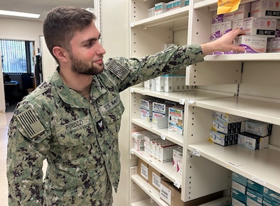 Uniformed service member stands in pharmacy and reaches for to high shelf for box of medication.