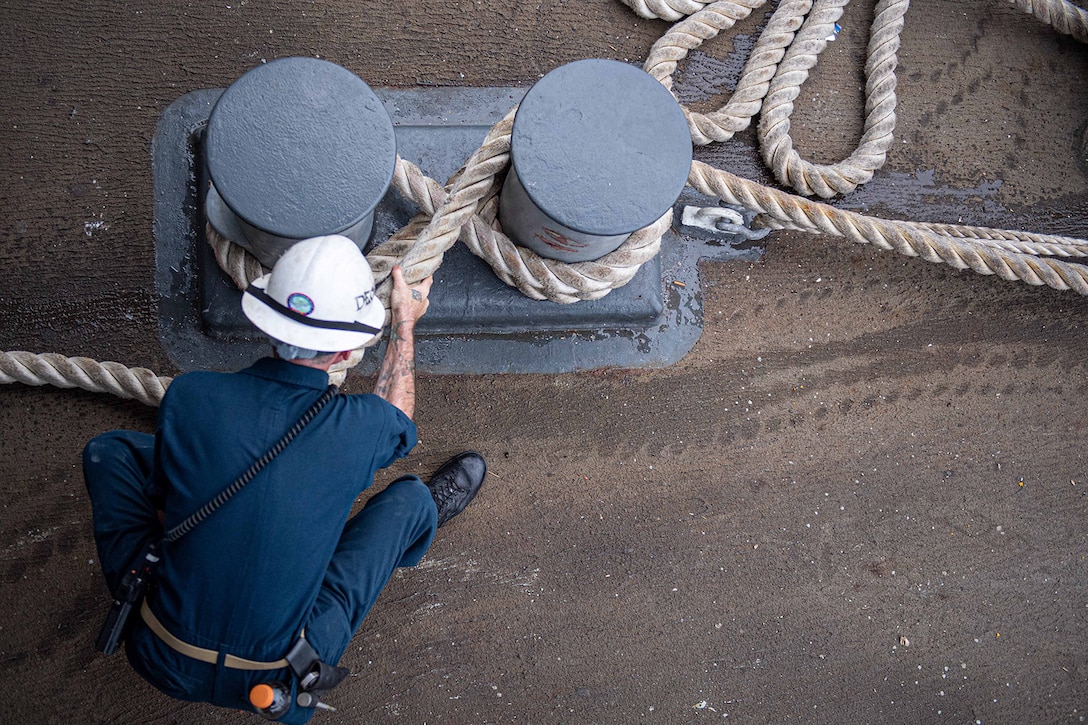 Overhead view of a sailor tying a mooring line onto a ship’s bitts.