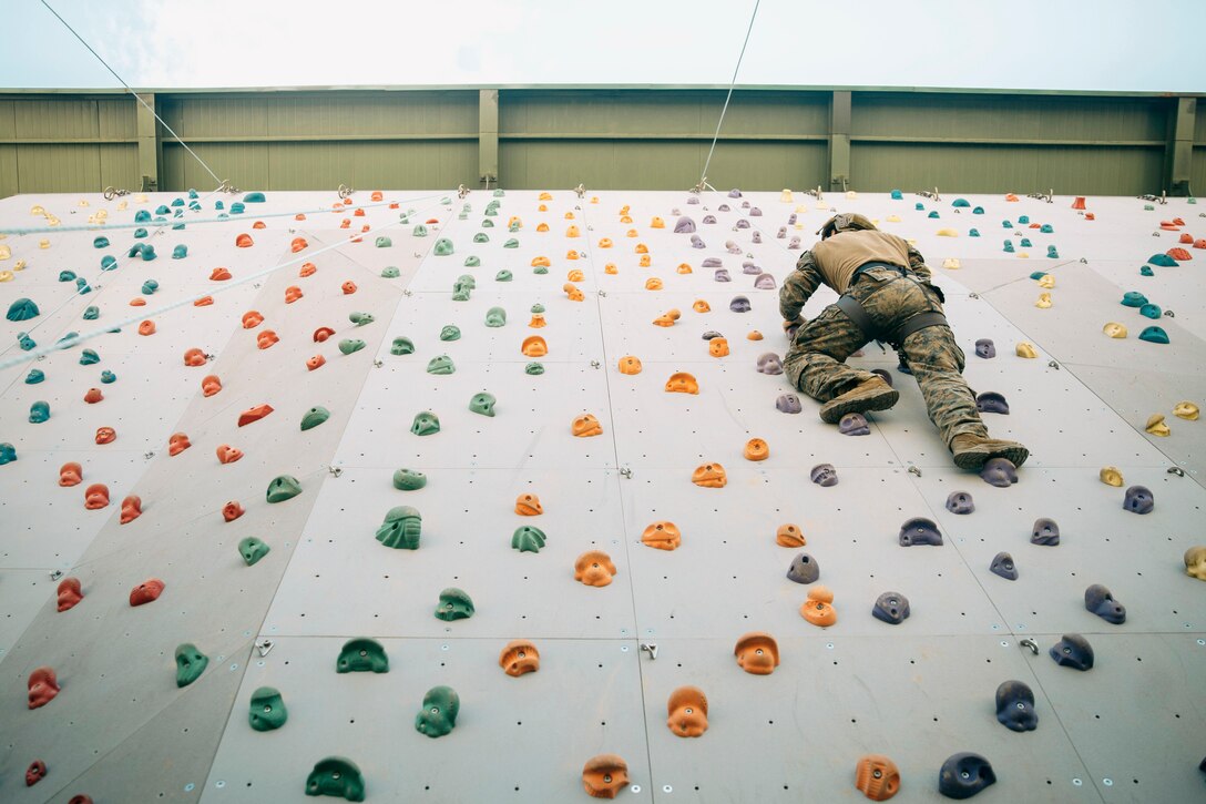 A Marine wearing a harness and a helmet climbs up a rock wall as seen from below.