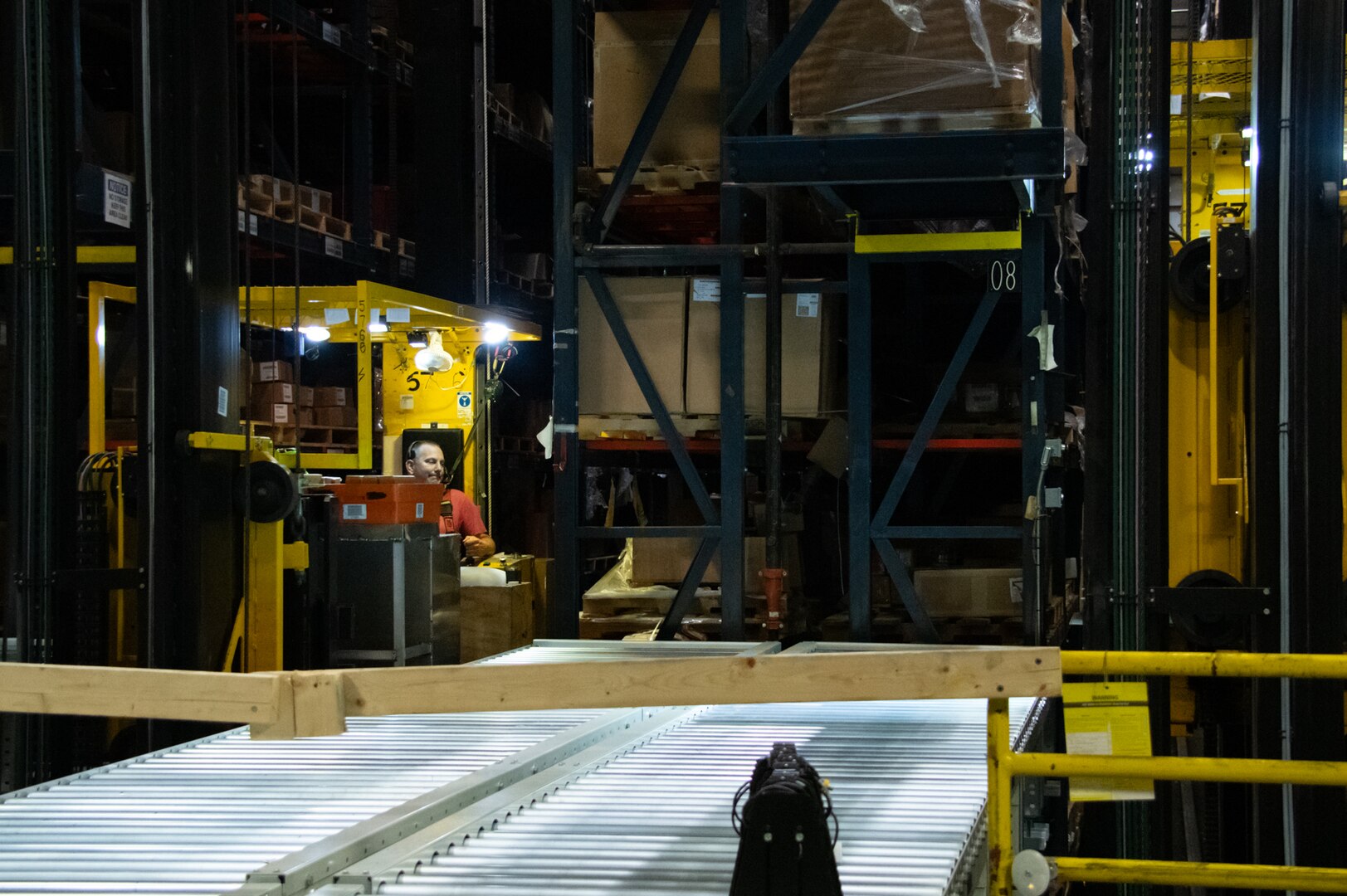 A man operates a large yellow crane amongst large storage shelves in a warehouse.