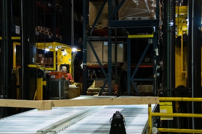 A man operates a large yellow crane amongst large storage shelves in a warehouse.