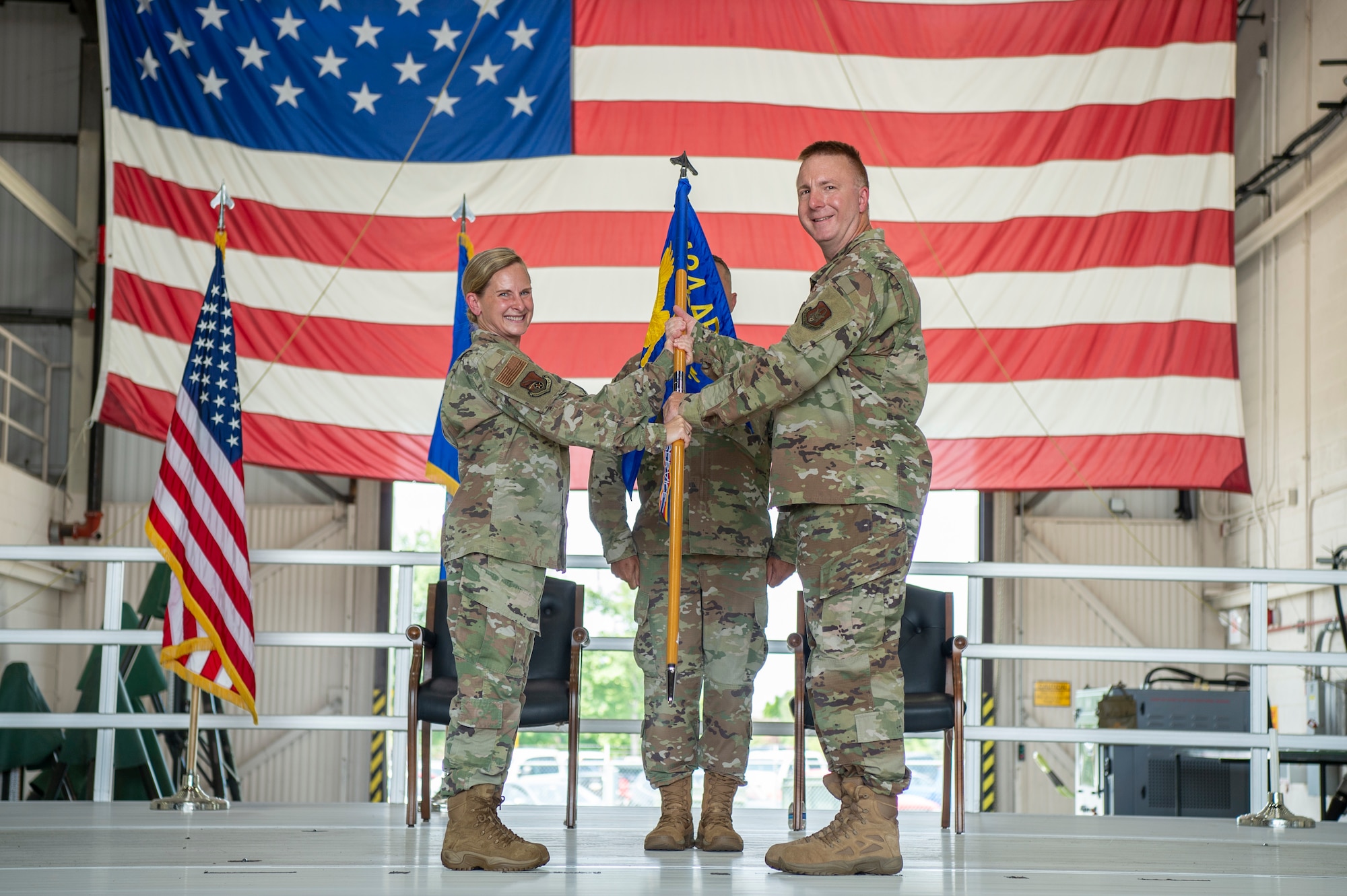 Two uniformed military members pose for a photo while holding a guidon. In the background, a third military member and giant U.S. flag.