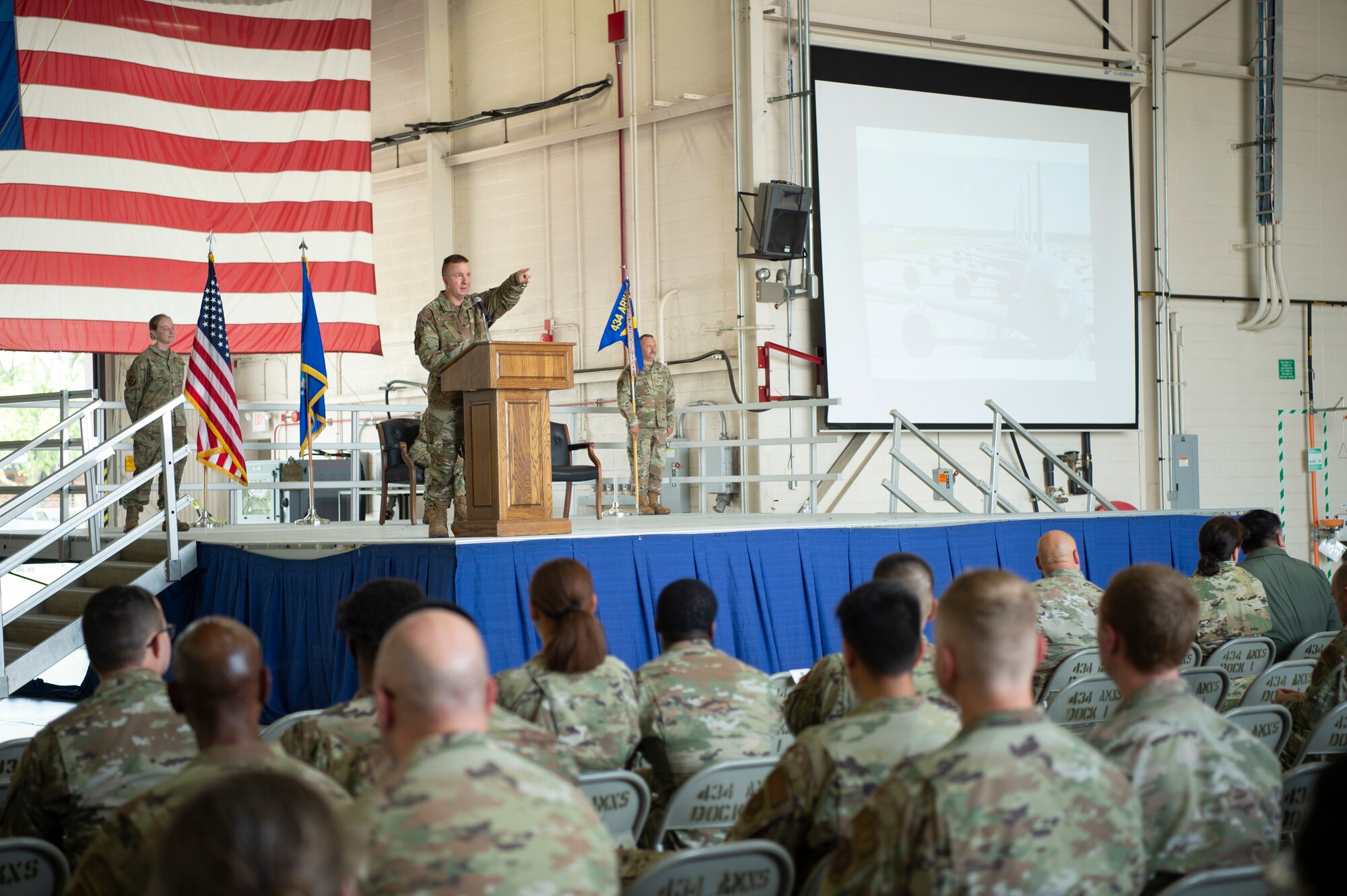 A uniformed military member is seen speaking on-stage, behind a podium. In front of him, a crowd of seated military members.
