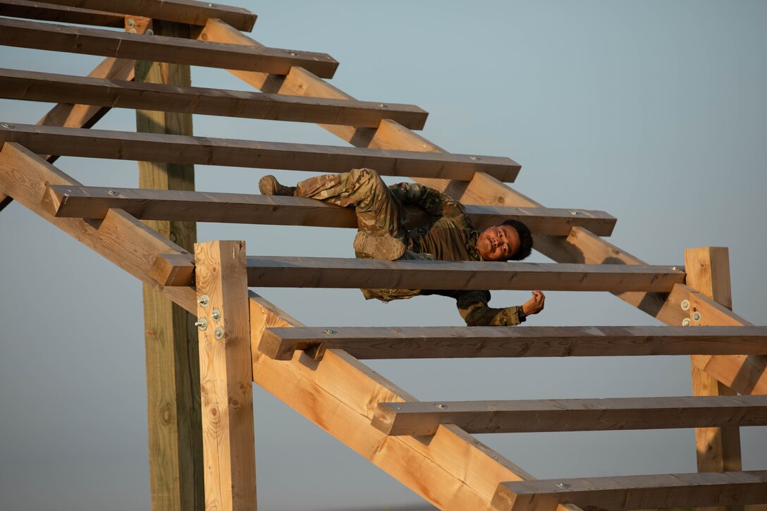 A soldier grimaces while using their arms and legs to cling to a large wooden angled ladder.