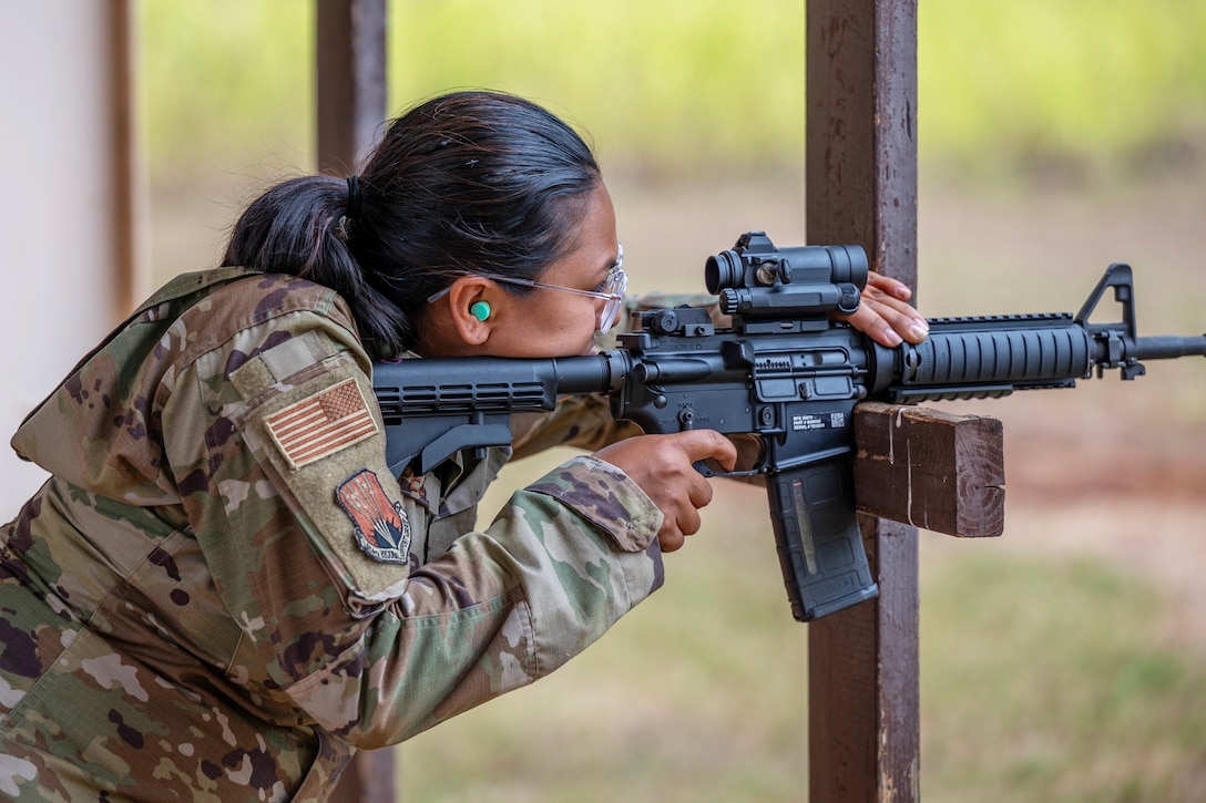 An airman wearing ear plugs balances a machine gun on a wooden beam while firing it during a weapons training exercise.