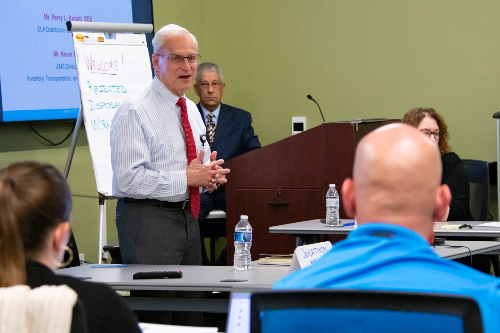 A man wearing a red tie speaks to a room full of people