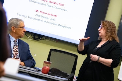 A woman speaks to a man who is sitting at a table. There is a display screen behind them.