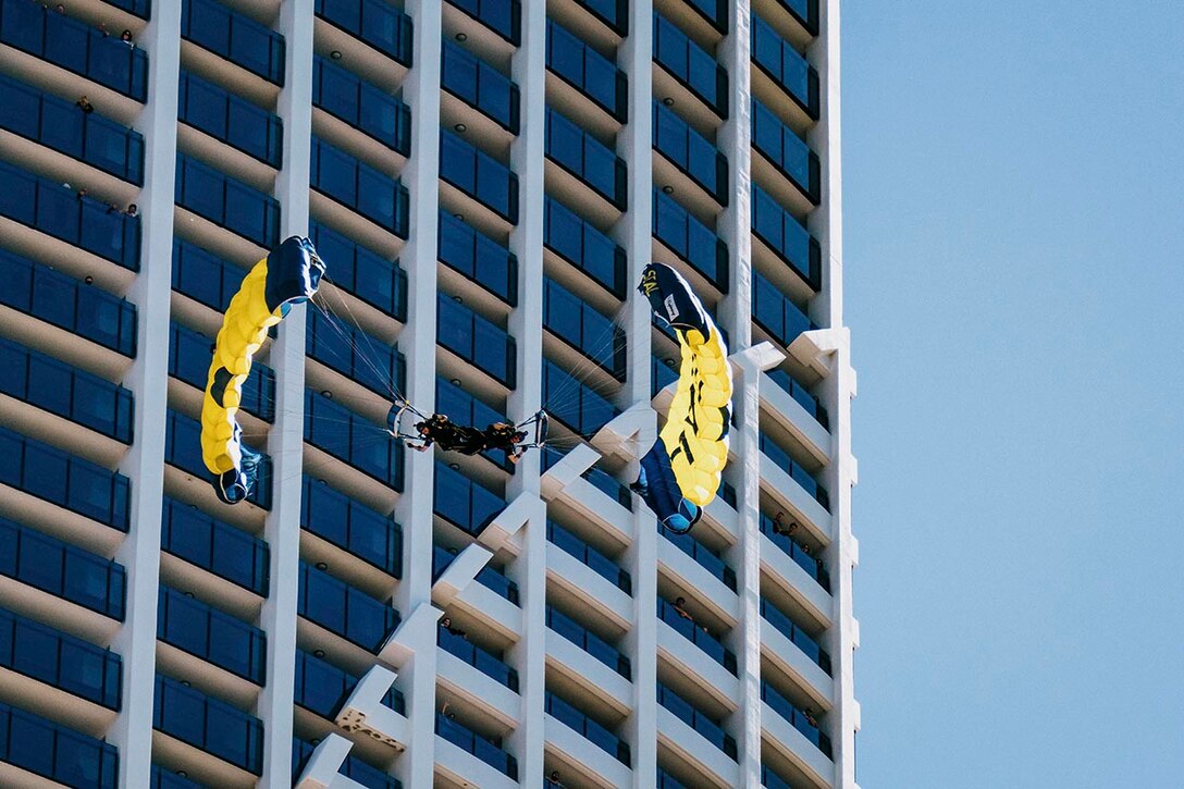 Two military parachute team members descend during daylight with a high-rise building in the background.