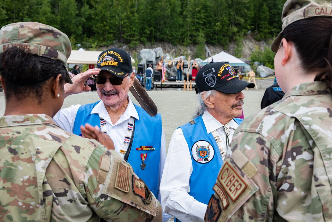 A veteran salutes an airman as another veteran and airman stand nearby. The veterans are wearing blue vests with insignia and baseball caps.