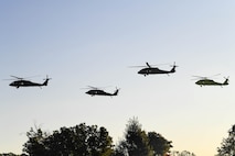 Four Army helicopters fly over a tree line