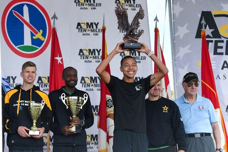 The men's division winners of the 39th annual Army-Ten Miler race hold their trophies in front of them on a stage as they are recognized. In the background are Army and race representatives dressed in casual attire.