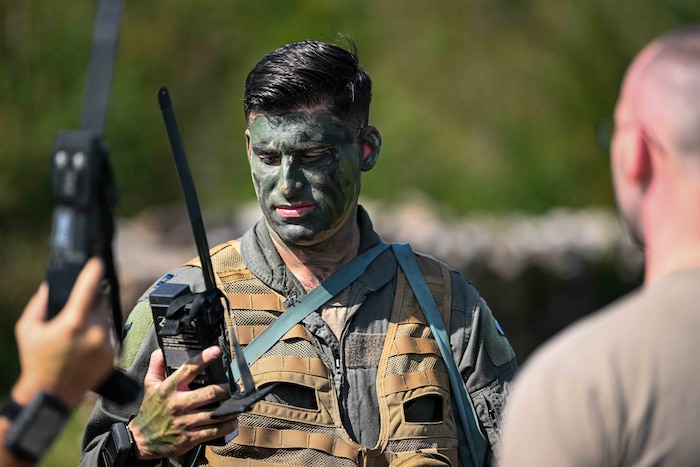 A U.S. Air Force pilot wearing a combat vest and camouflage face and hand paint looks a radio he's holding.
