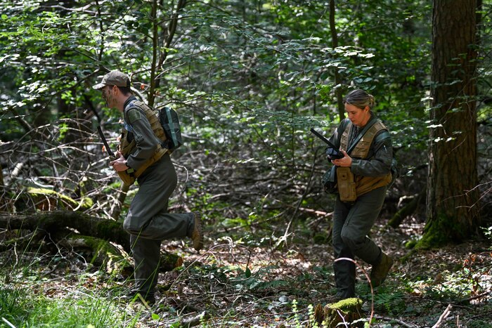 A male and female U.S. Air Force pilot wearing combat vests hold radios and walk through the woods.