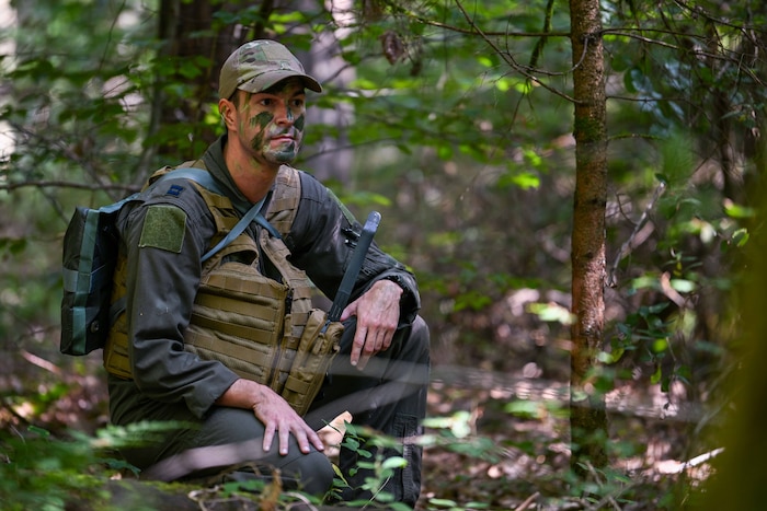 A U.S. Air Force pilot wearing a camouflage hat and face paint and a combat vest takes a knee and looks around in the woods.