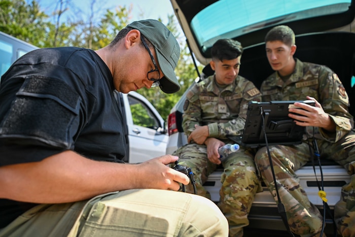 A civilian and two U.S. Air Force Airmen sit behind a car and look a remote controller and a monitor.