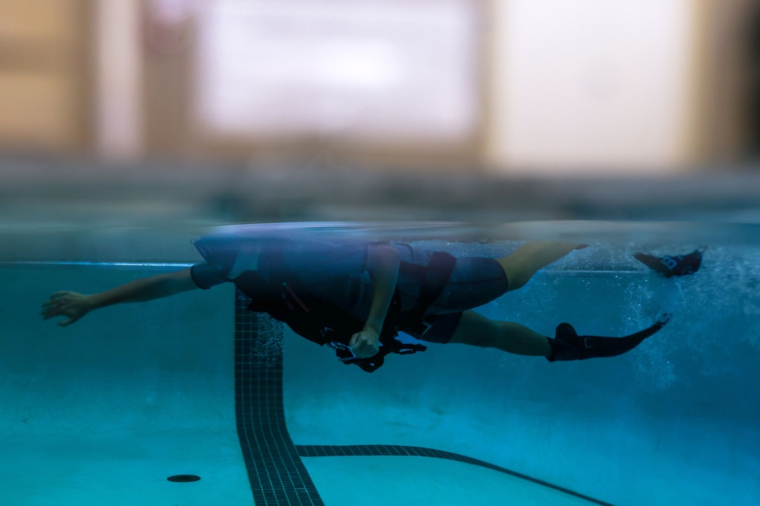 A sailor wearing goggles and fins swims laps in a pool as seen from underwater.