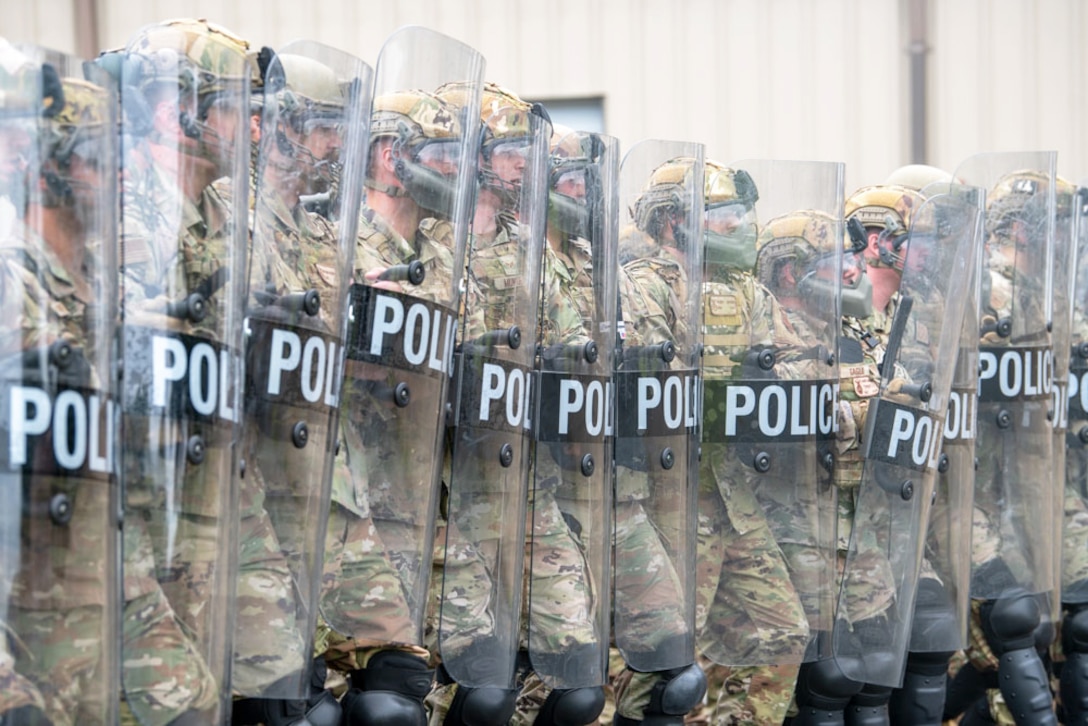 A dozen National Guardsmen wearing protective gear stand in formation while holding police shields outside of a building.