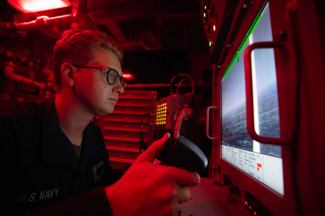 A sailor looks at a monitor while operating a console in a control center illuminated by red light.