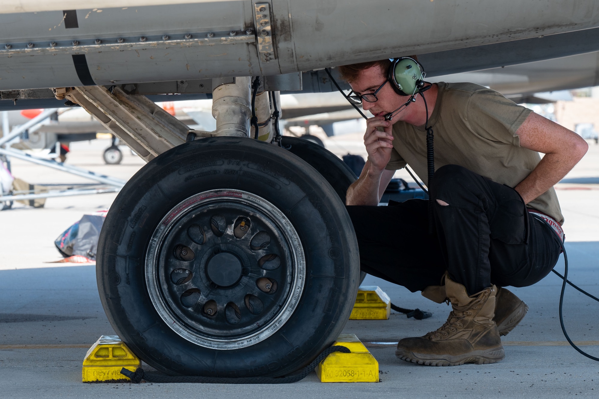 A man kneeling by a landing gear speaks into a microphone attached to his headset.