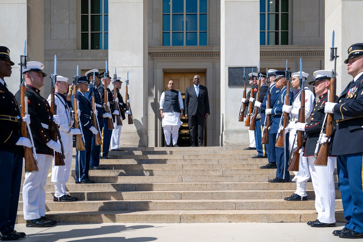 Two people in civilian clothing stand atop a staircase flanked by members of the military in dress uniform.