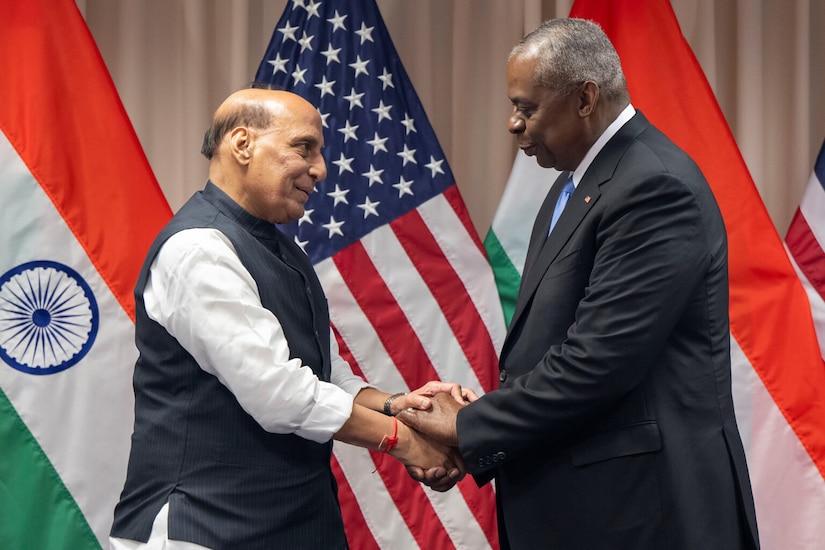 Two people in business attire shake hands in front of U.S. and Indian flags.