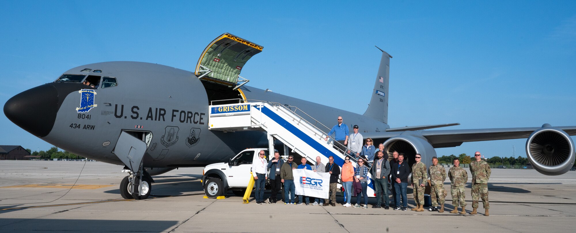 A group of men and women, including some uniformed service members, pose for a photo in front of a military aircraft.