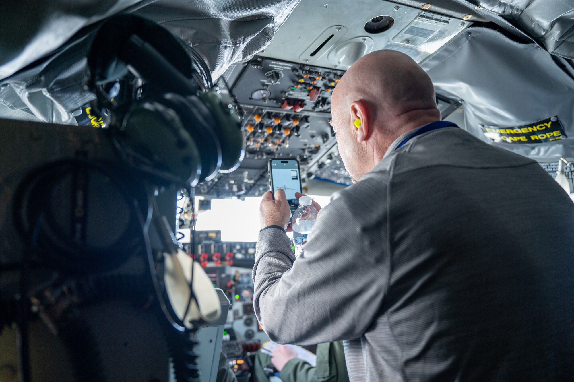 A man in civilian clothing takes a photo while in the cockpit of a military aircraft. Visible on his phone screen, the live image of another military aircraft. Both aircraft are KC-135s.