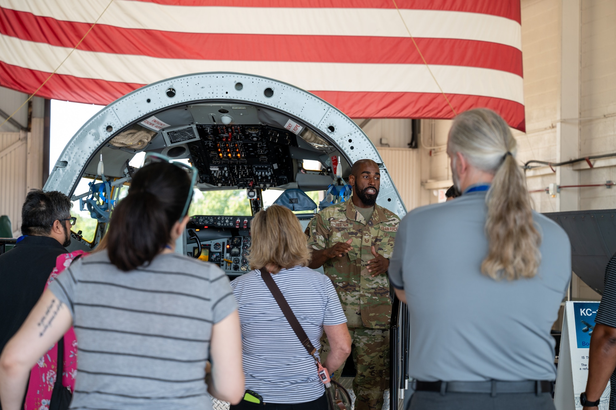 A man in a military uniform talks to civilians. In the far background, the bottom of a giant American flag. The man is standing in front of a cockpit display of a military aircraft.