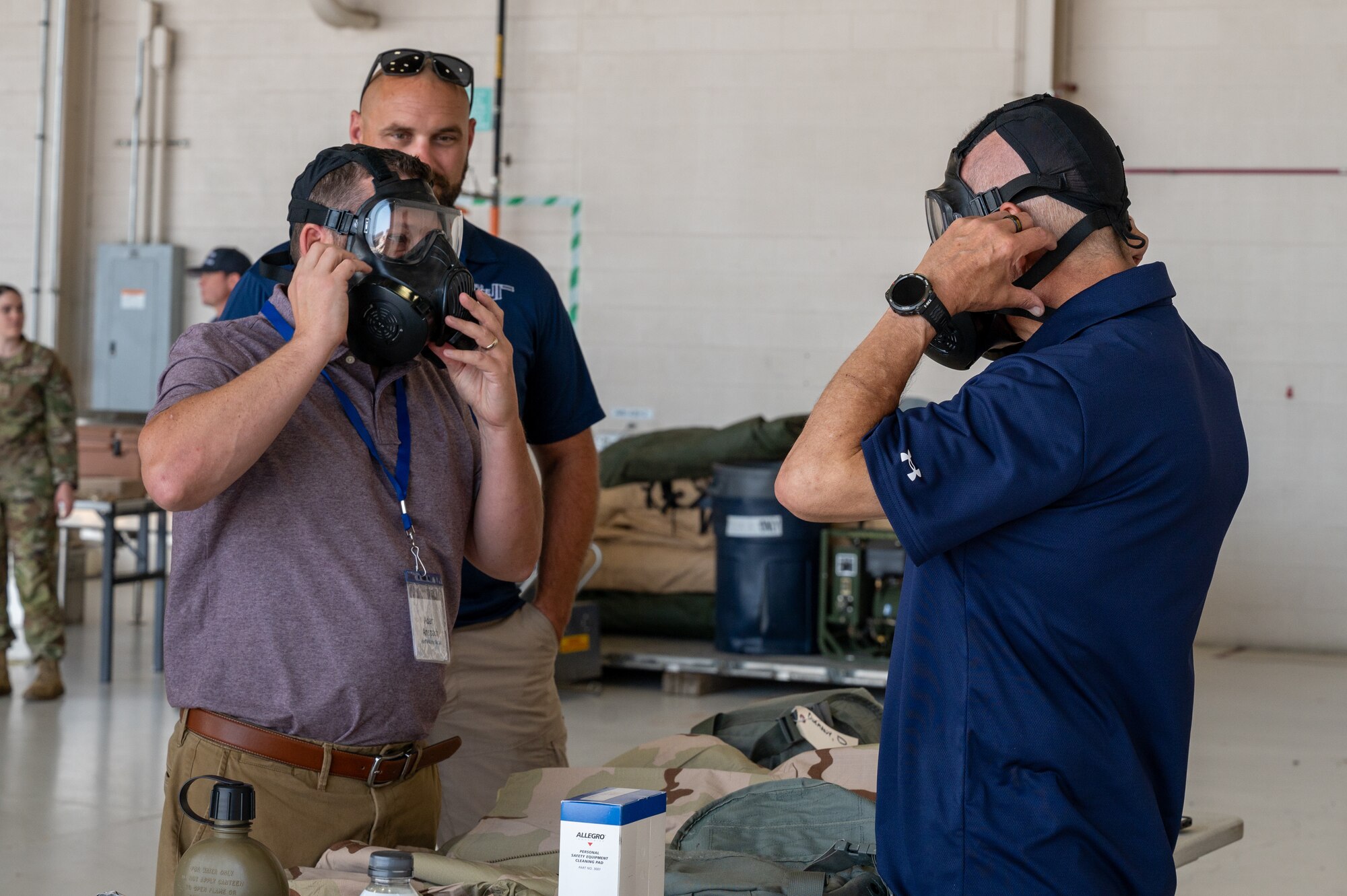 Two men stand face-to-face as they pull military-grade gas masks over their faces. A third man observes in the background.