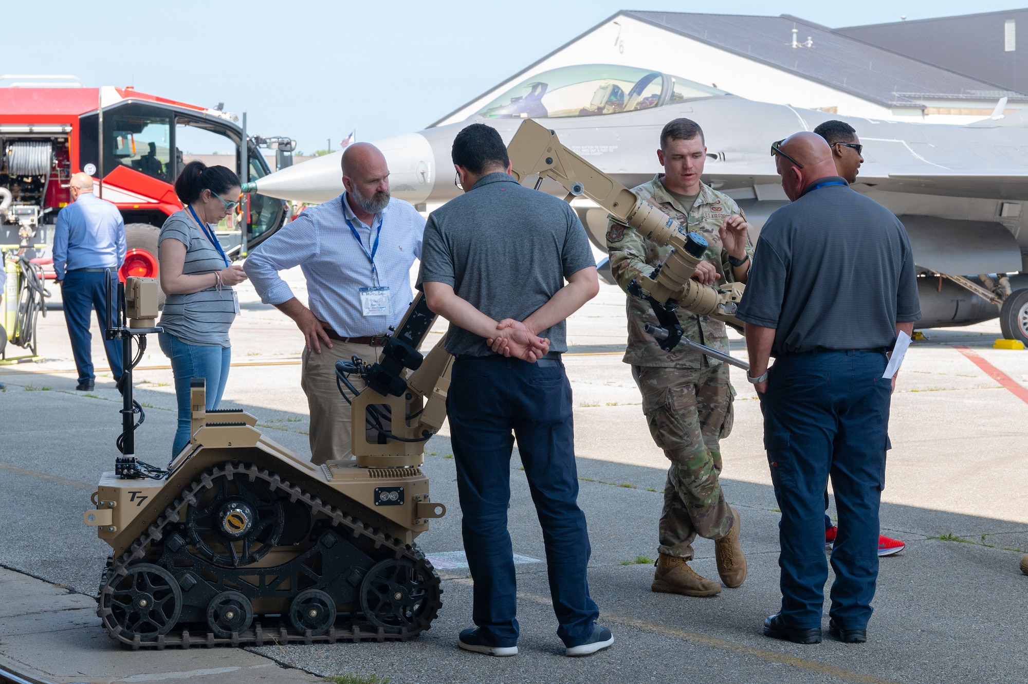 Men and women talk to one another while standing around a military robot. A fighter jet is visible in the background.