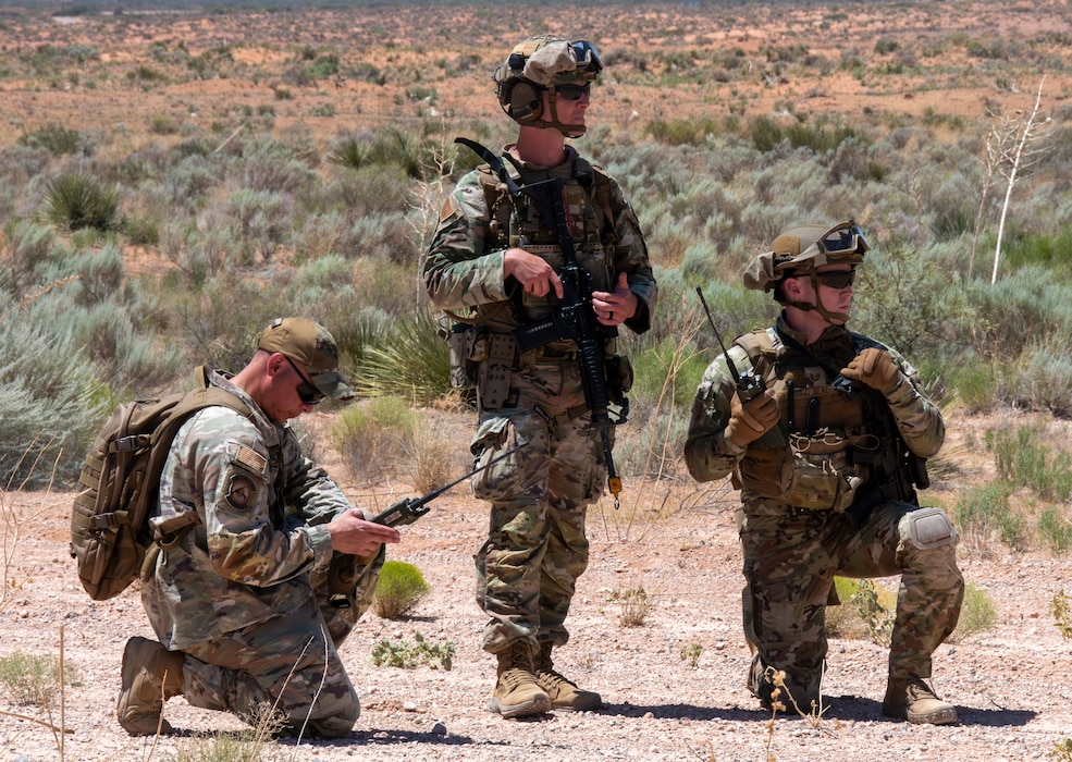 Three Airmen, two kneeling and one standing, in the desert awaiting instructions during a training exercise