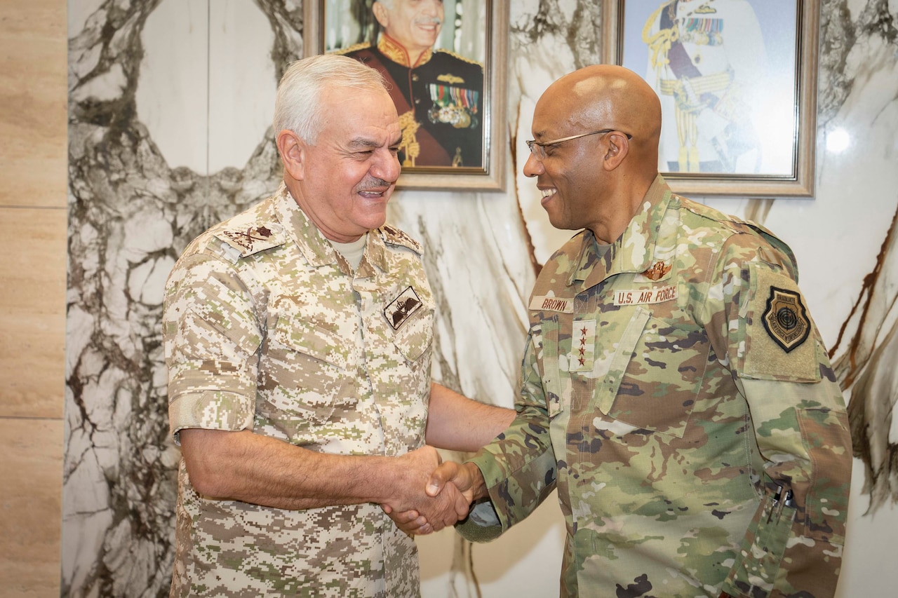 Two service members in camouflage uniforms are smiling and shaking hands while looking at each other.