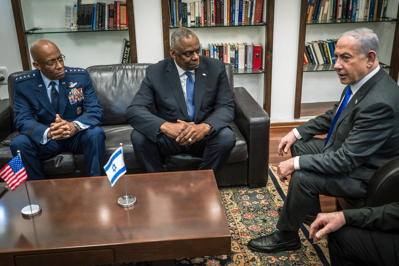 A service member and two civilians sit around a wooden table that has one small U.S. flag and one small Israeli flag atop it.