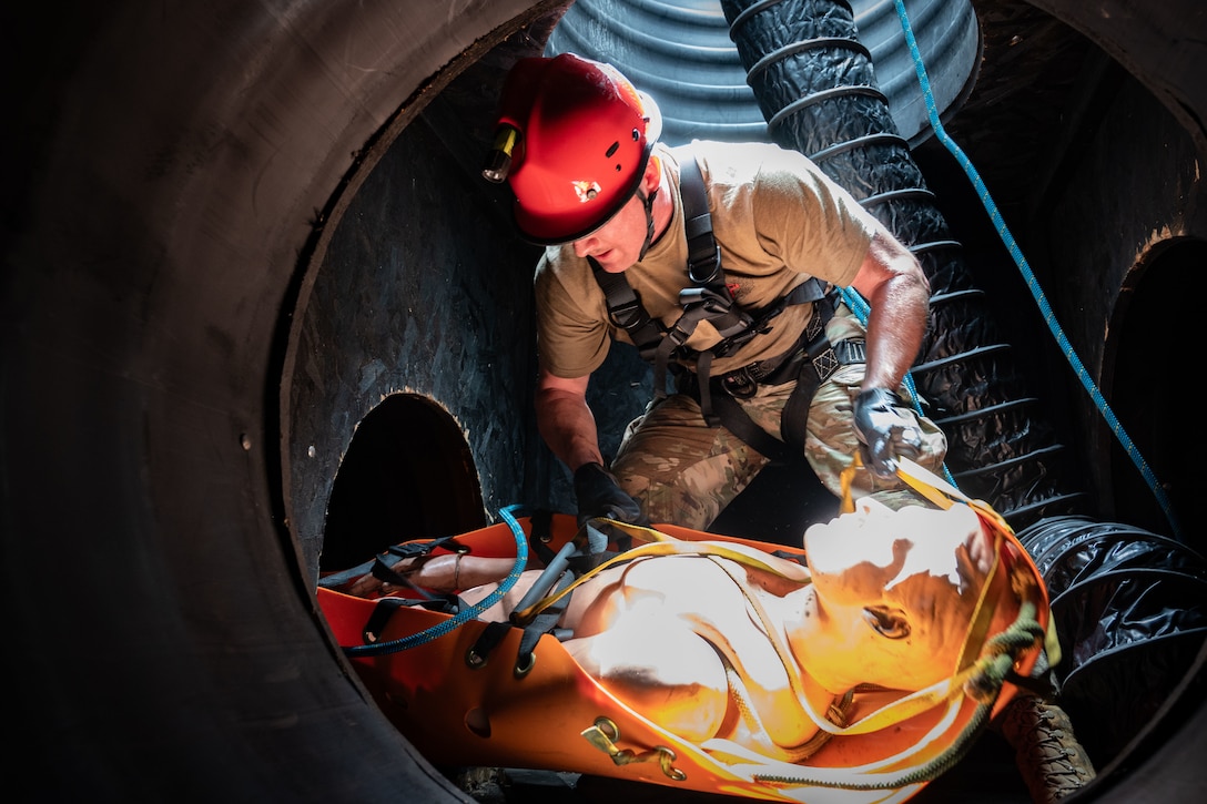 An airman wearing a safety helmet with a headlamp bends over a litter carrying a training mannequin in an underground space.