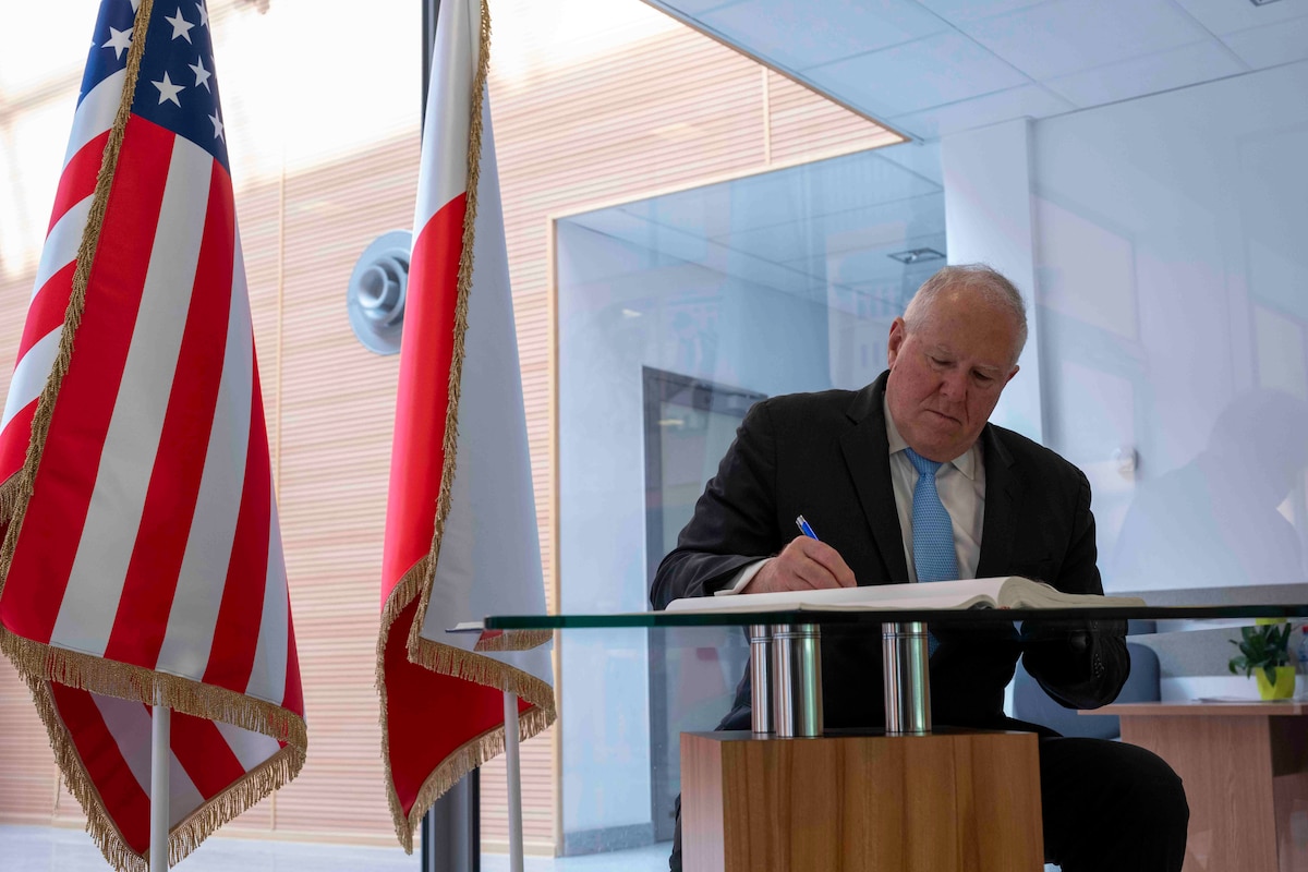 A man in a business suit signs a book at a table to the right of an American and Polish flag.