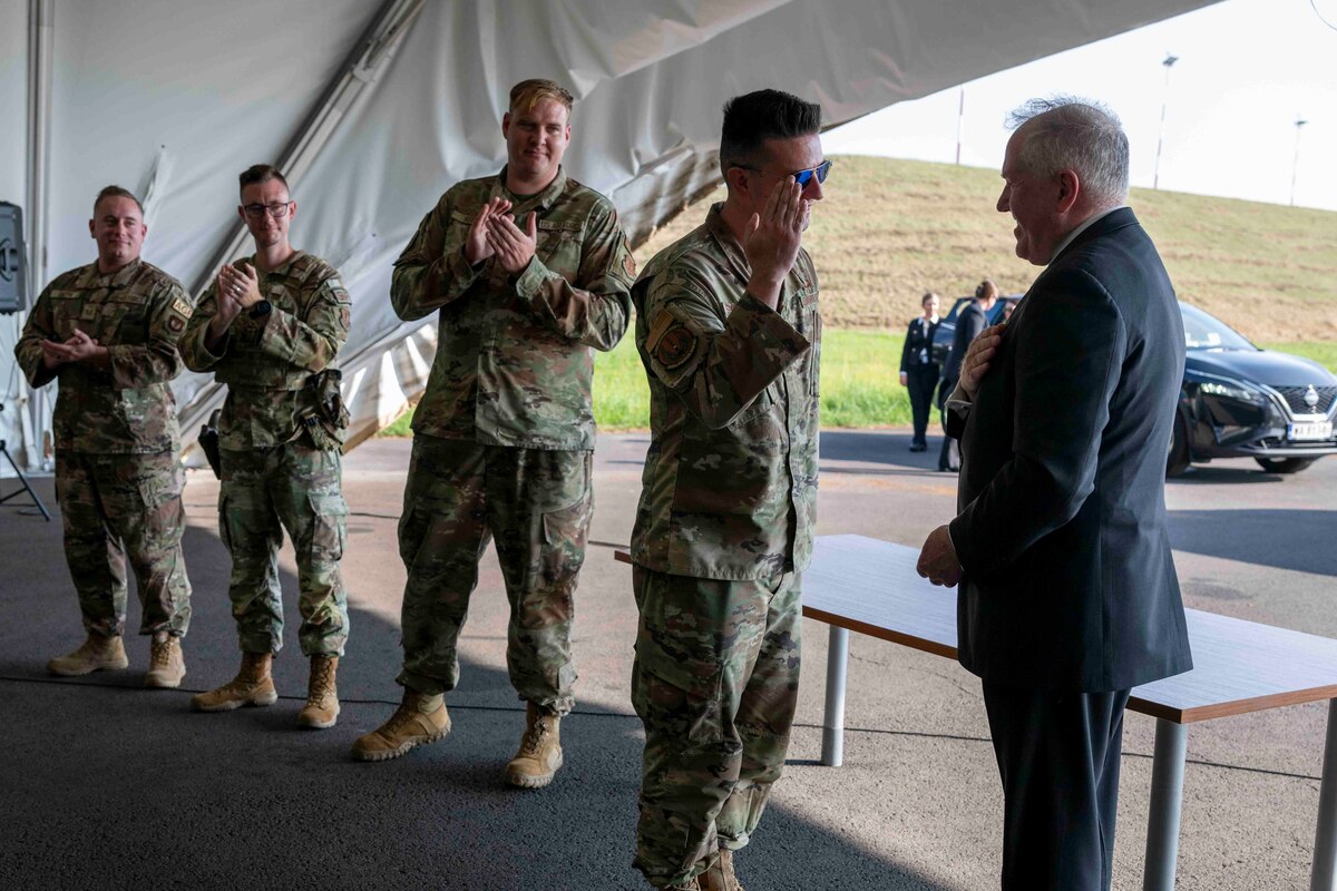 A U.S. Air Force Airmen salutes a man in a suit while other Airmen to his left in the background clap underneath a tent.