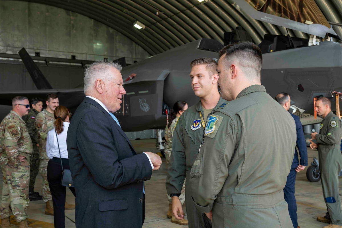 A man in a business suit talks with two U.S. Air Force pilots in front of a fighter jet in a hangar.