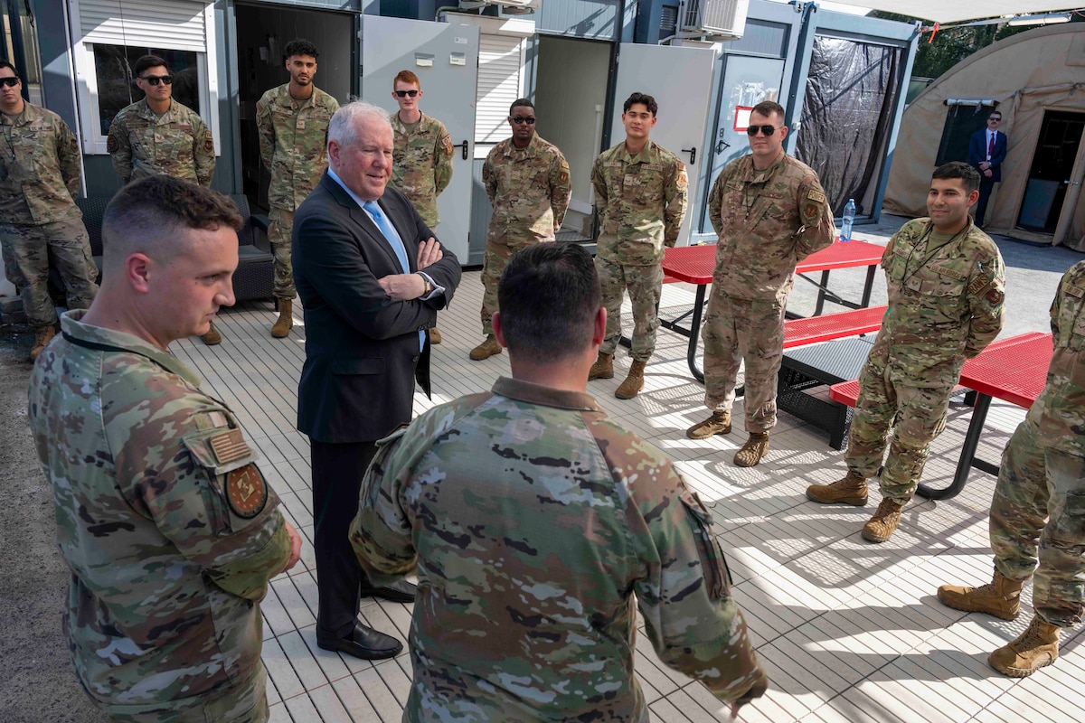 A man in a suit stands with his arms folded surrounded by a circle of U.S. Air Force Airmen.